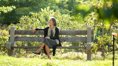 Virginia Chang, instructor for UVM's End-of-Life Doula professional certificate, sitting on a park bench with lush green scenery behind the bench.