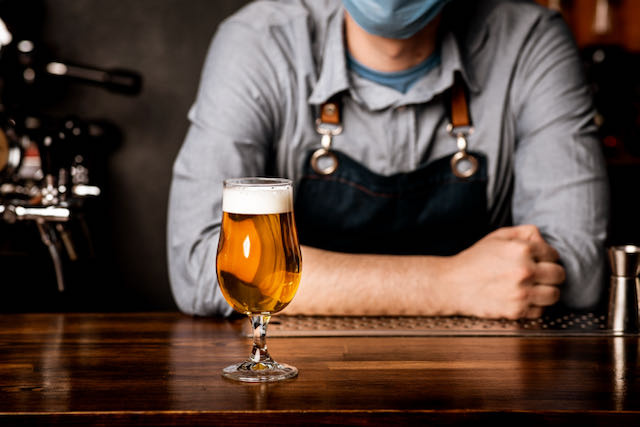 beer glass on counter with bartender in the background wearing a mask during covid 19