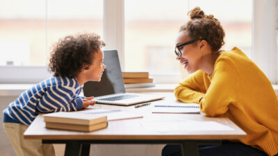 Young happy afro american woman mom talking with little kid son while working on laptop online at home