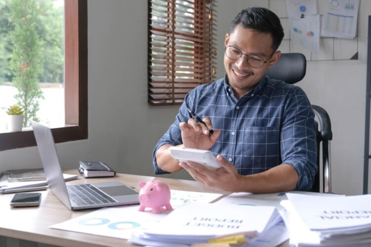 Portrait of an Asian businessman using a calculator to calculate his savings from SME operations, with a pink piggy bank as keep money concept.