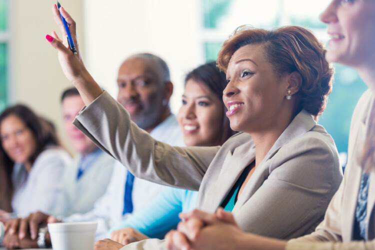 Mid adult African American businesswoman is smiling and raising her hand. She is answering a question during a business conference or job training seminar. Professional woman is wearing business clothing. She is sitting with diverse colleagues.