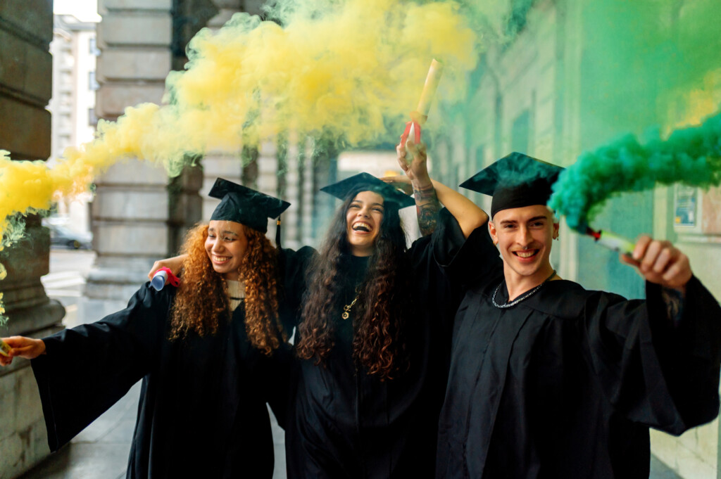 Graduates in gowns smiling and celebrating with green and gold smoke flares.