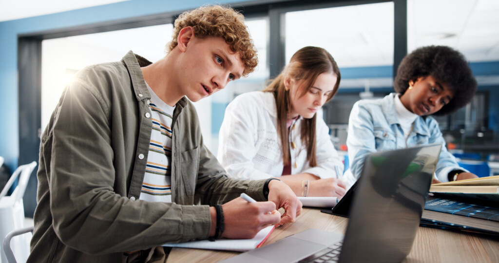 Group of students studying at a table with a boy in the foreground looking at laptop while writing.