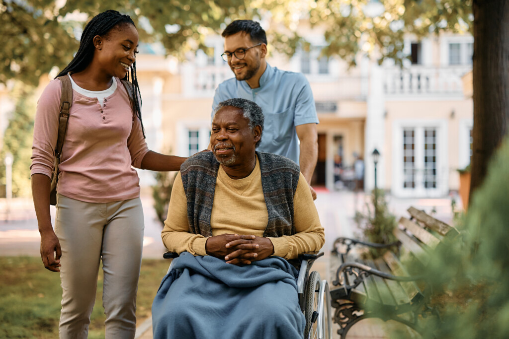 End-of-Life Doula Certificate portfolio at University of Vermont. Happy senior man in wheelchair talking to his daughter who is visiting him in nursing home, with end-of-life doula pushing wheelchair.