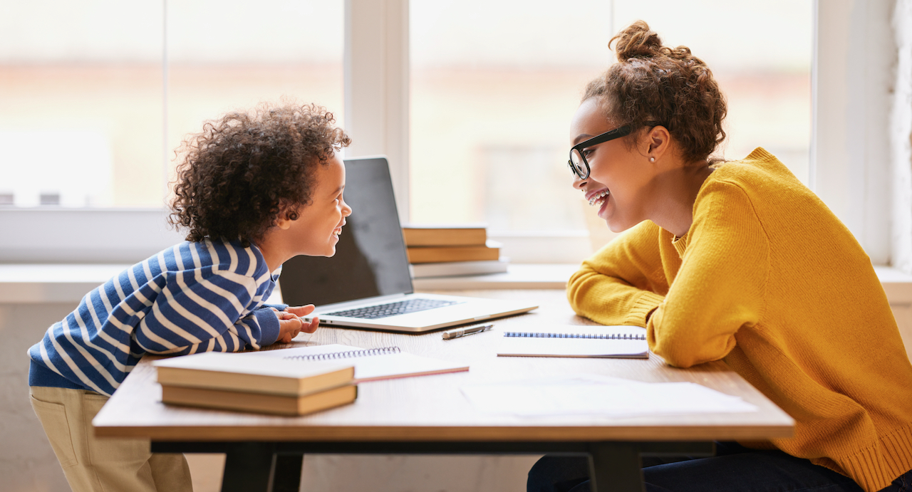 Cute boy with curly hair talking with smiling mom while she working remotely on laptop from home. Childcare concept
