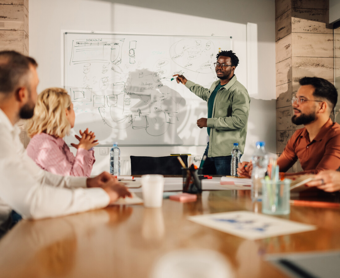 Group of professionals sitting around a conference room table in an office looking at a man presenting at a white board in the front of the room.