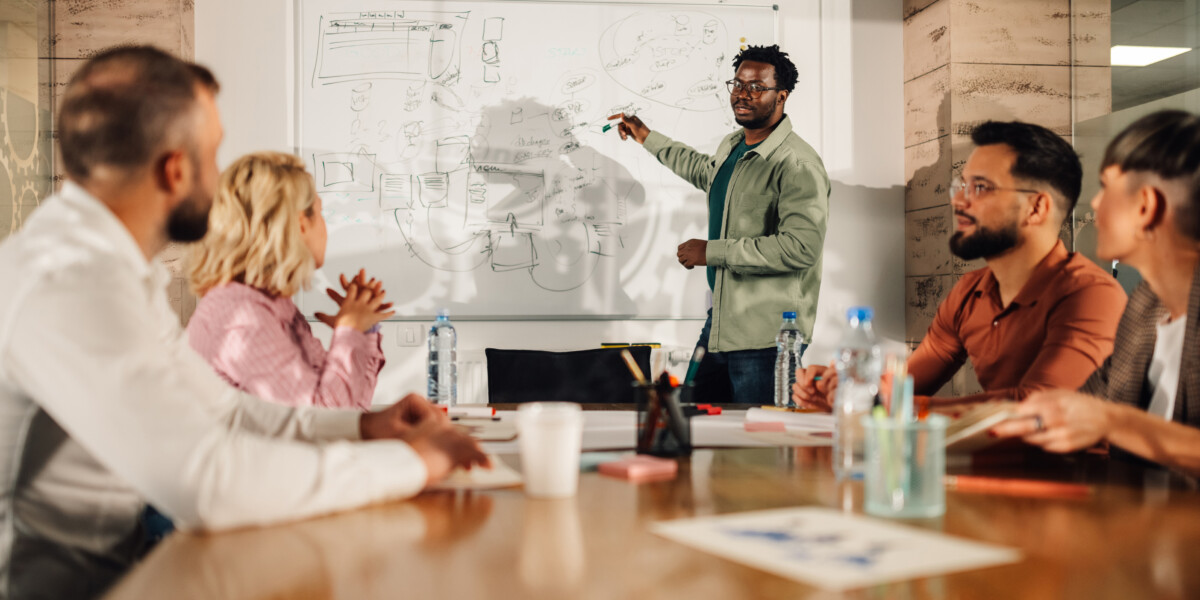 Group of professionals sitting around a conference room table in an office looking at a man presenting at a white board in the front of the room.
