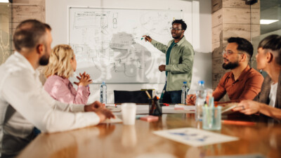 Group of professionals sitting around a conference room table in an office looking at a man presenting at a white board in the front of the room.