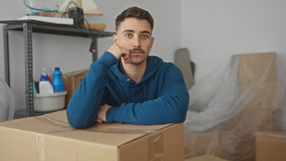 Person in a blue hoodie leaning on cardboard box in new apartment surrounded by unpacked item.