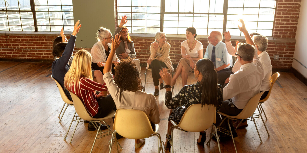Group of adult learners and students sitting in a circle, some with hands raised, discussing topics in UVM's End-of-Life Care portfolio of certificates.