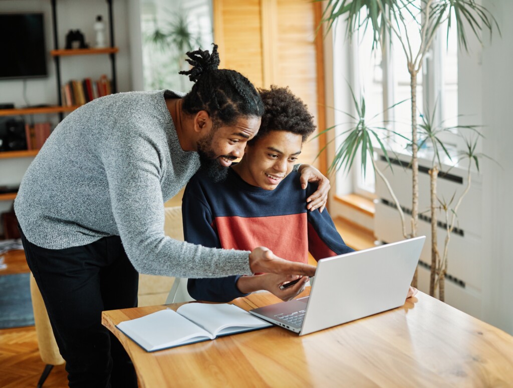 High school student and guardian are looking at a laptop on a table, smiling and working together.