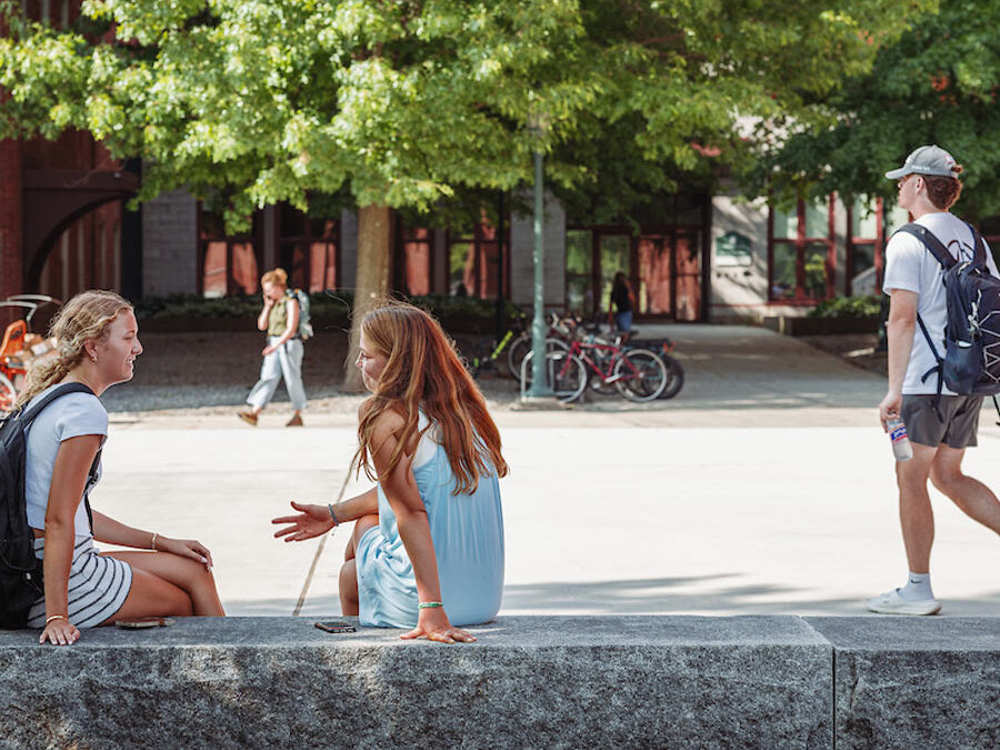 Students sit on a bench and walking in the background during a summer day on campus at University of Vermont.