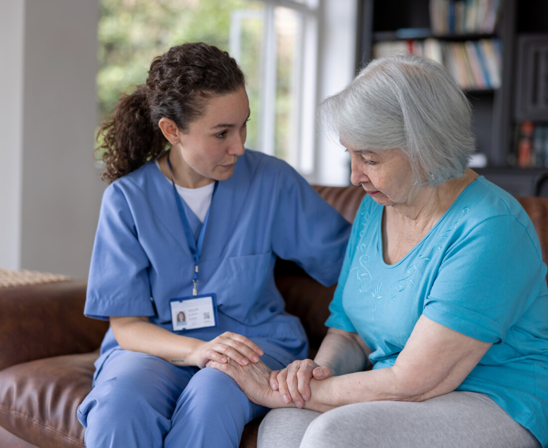 A healthcare worker in blue scrubs, wearing an ID badge, is sitting on a brown couch comforting an older person with gray hair, who is wearing a light blue shirt. They are in a room with a bookshelf and windows in the background.