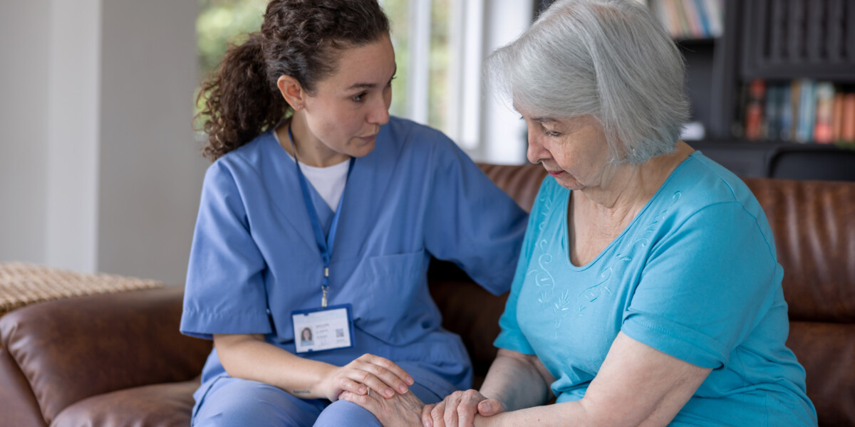 A healthcare worker in blue scrubs, wearing an ID badge, is sitting on a brown couch comforting an older person with gray hair, who is wearing a light blue shirt. They are in a room with a bookshelf and windows in the background.