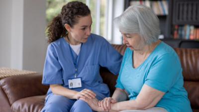 A healthcare worker in blue scrubs, wearing an ID badge, is sitting on a brown couch comforting an older person with gray hair, who is wearing a light blue shirt. They are in a room with a bookshelf and windows in the background.