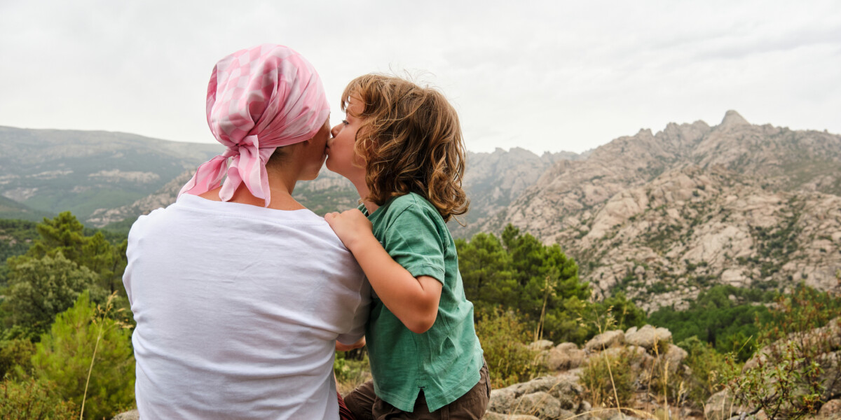 Two people kissing outdoors with a rocky mountain range in the background, under a cloudy sky.