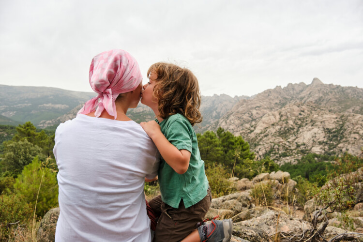 Two people kissing outdoors with a rocky mountain range in the background, under a cloudy sky.