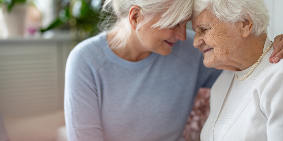 Two elderly women share a tender moment, foreheads touching and smiling, indoors near soft natural light.