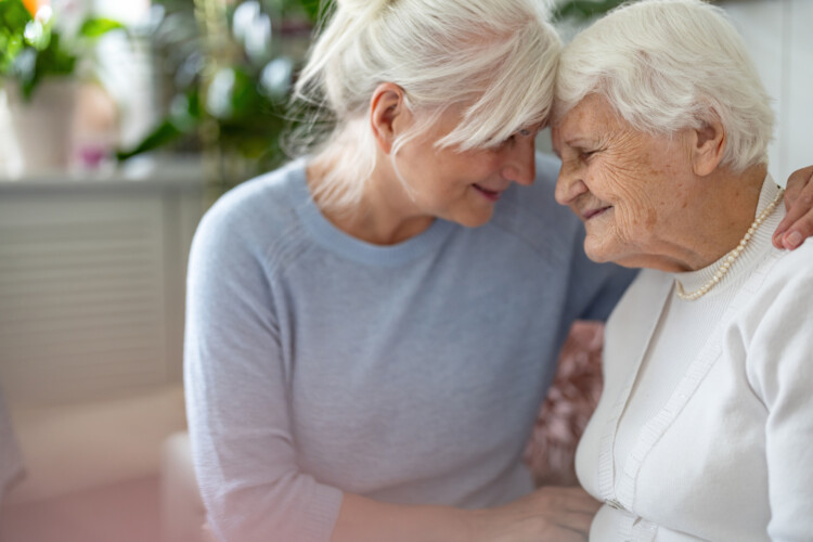 Two elderly women share a tender moment, foreheads touching and smiling, indoors near soft natural light.