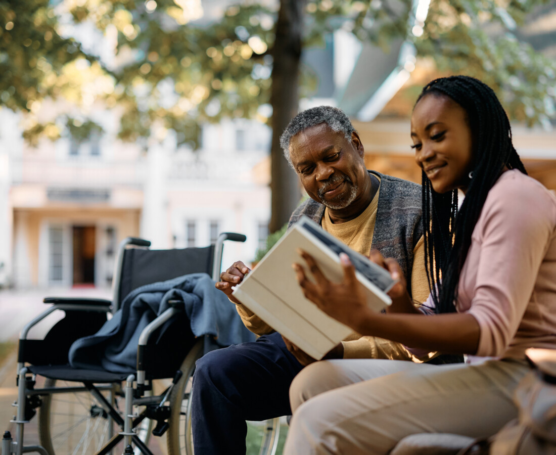 Two people outdoors in a park, an older man in a wheelchair and a younger woman reading a book together on a bench.