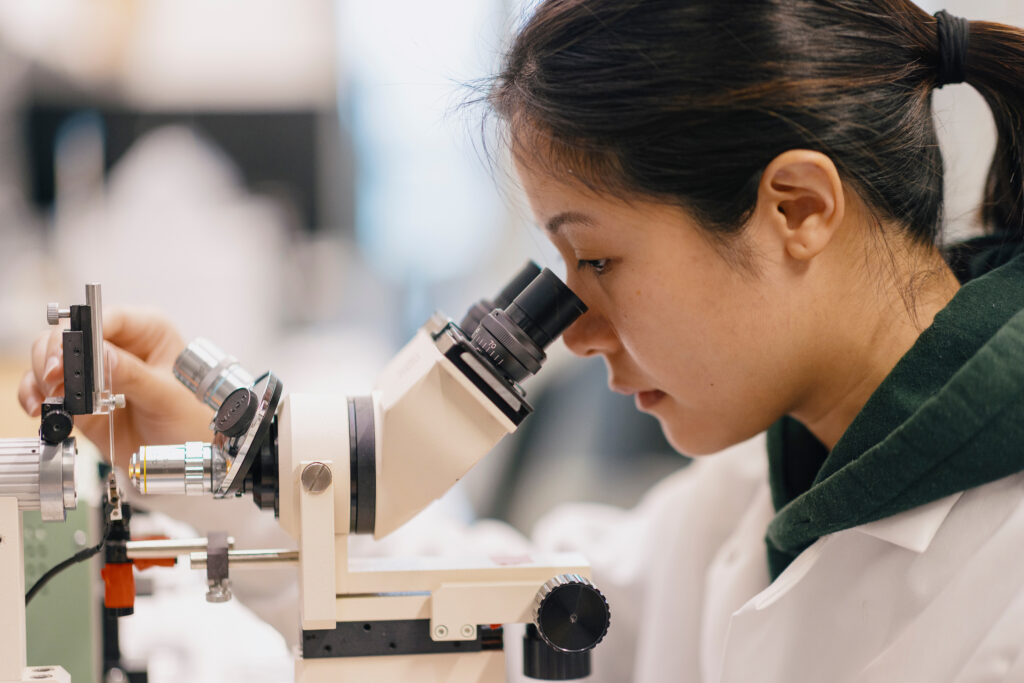 Firestone-Medical-Research-Lab_LCOM_R1-Press_Photog-Joshua-Defibaugh_File-name_Defibaugh_R1_Press-0646 - UVM Professional and Continuing Education Woman in a lab coat looking into a microscope, adjusting the stage in a bright lab setting