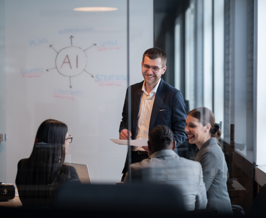Businessman in a dark suit presenting to colleagues in a glass-walled meeting room, with an AI diagram on the whiteboard.