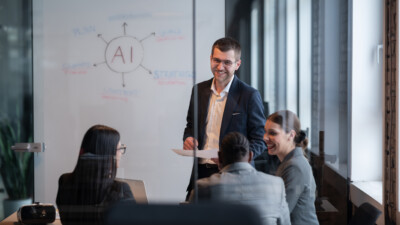 Businessman in a dark suit presenting to colleagues in a glass-walled meeting room, with an AI diagram on the whiteboard.