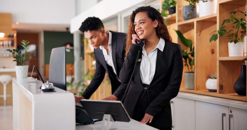 Hotel reception and call for booking consultant with workers customer care and hospitality enquiry for villa Staff service and question at lobby desk with conversation helping and management - UVM Professional and Continuing Education Receptionists at a modern front desk; woman on the phone smiling as a colleague assists nearby.