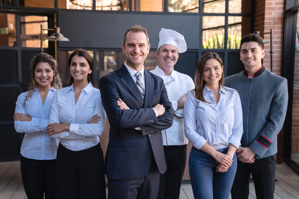 Cheerful latin american team of hotel staff all facing camera smiling - UVM Professional and Continuing Education Group of six hotel staff standing in a row and smiling in front of the lobby door.