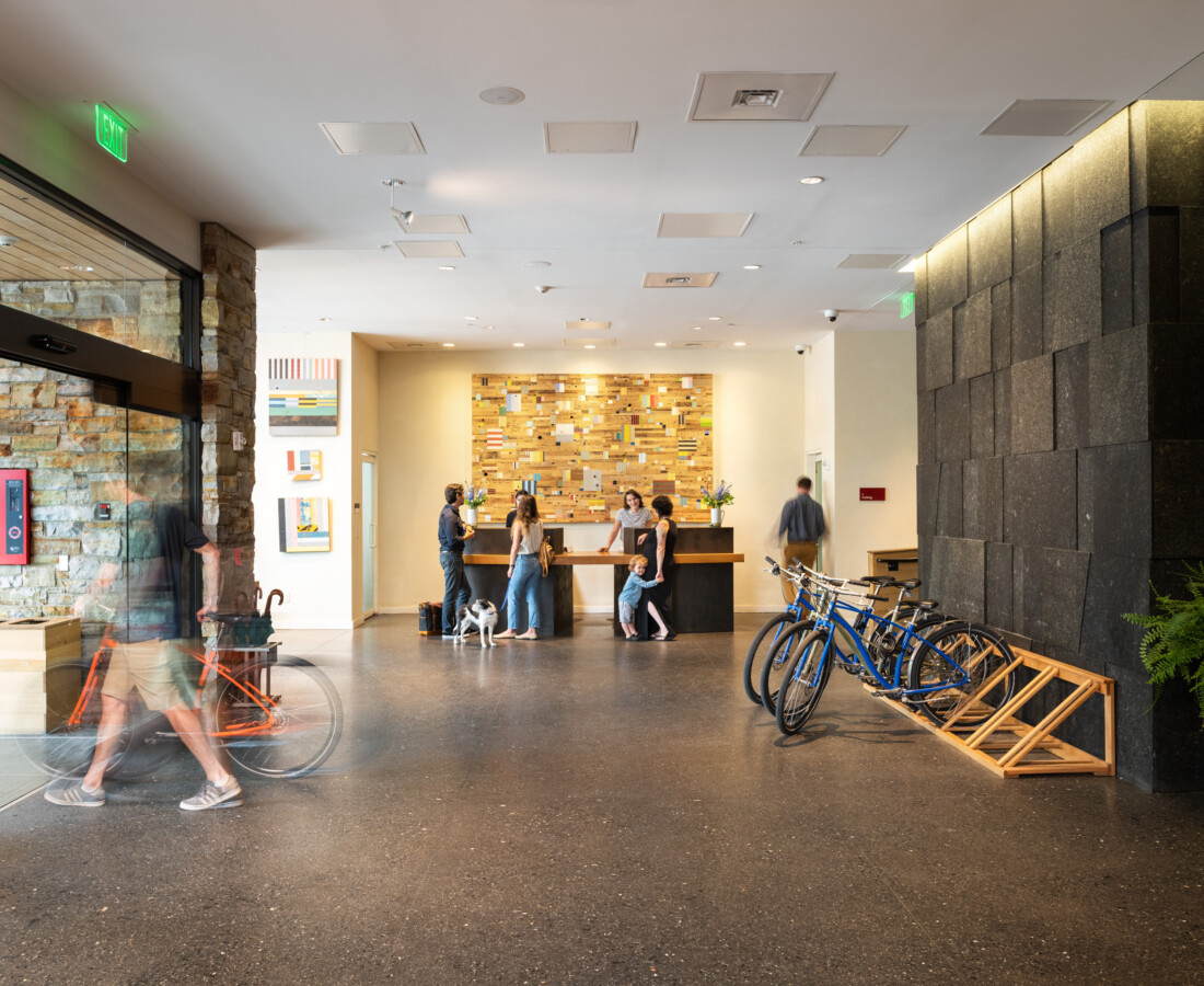 A bright lobby with a reception desk, several people chatting, bikes parked on the right and modern wood and stone decor in the background.