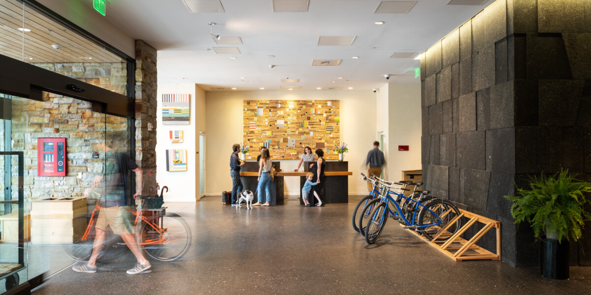 A bright lobby with a reception desk, several people chatting, bikes parked on the right and modern wood and stone decor in the background.