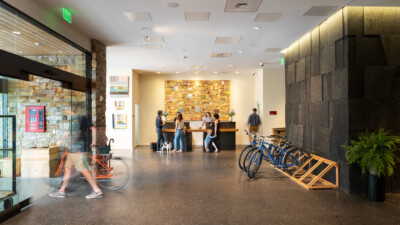 A bright lobby with a reception desk, several people chatting, bikes parked on the right and modern wood and stone decor in the background.