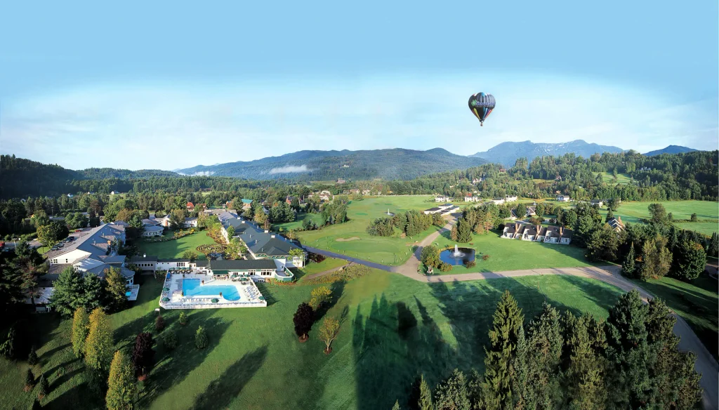 Stoweflake_Sales-Buyout-scaled-1 - UVM Professional and Continuing Education Aerial view of a resort town with houses, a pool, and manicured greens, as a colorful hot air balloon floats over distant hills.
